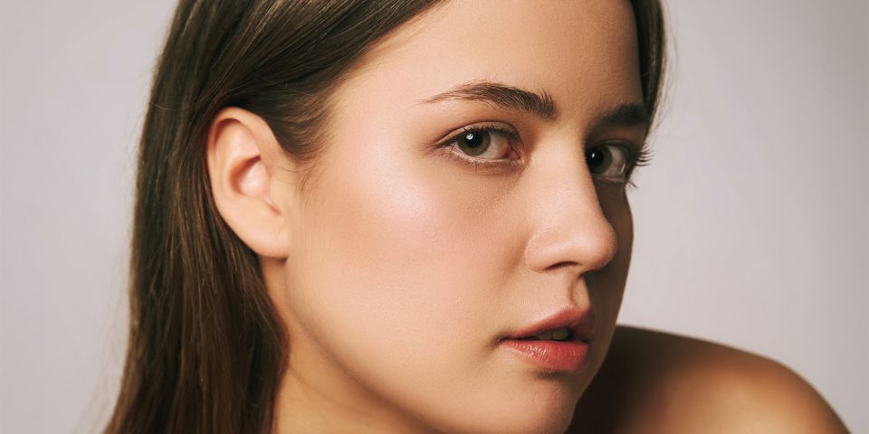 A young woman with natural makeup and long brown hair looking over her shoulder against a soft grey background
