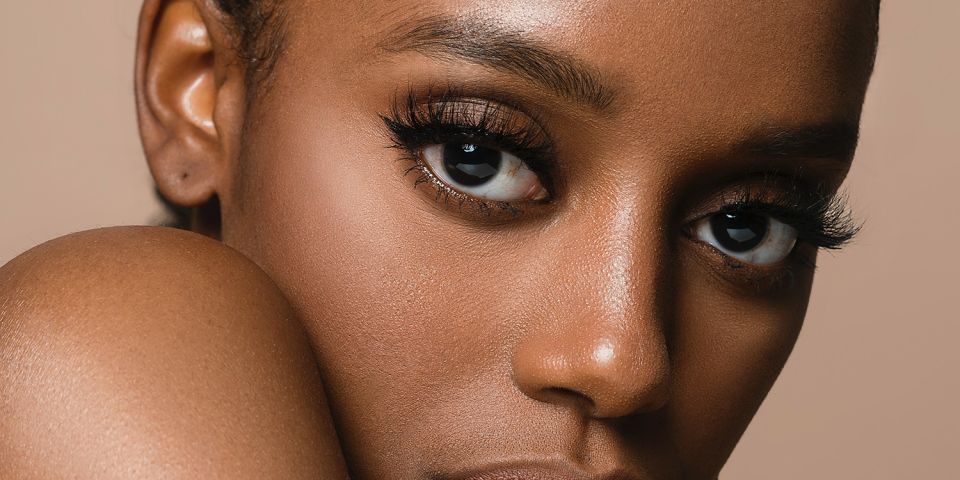 A close-up portrait of a woman with glowing, healthy skin resting her chin on her hand against a warm beige background